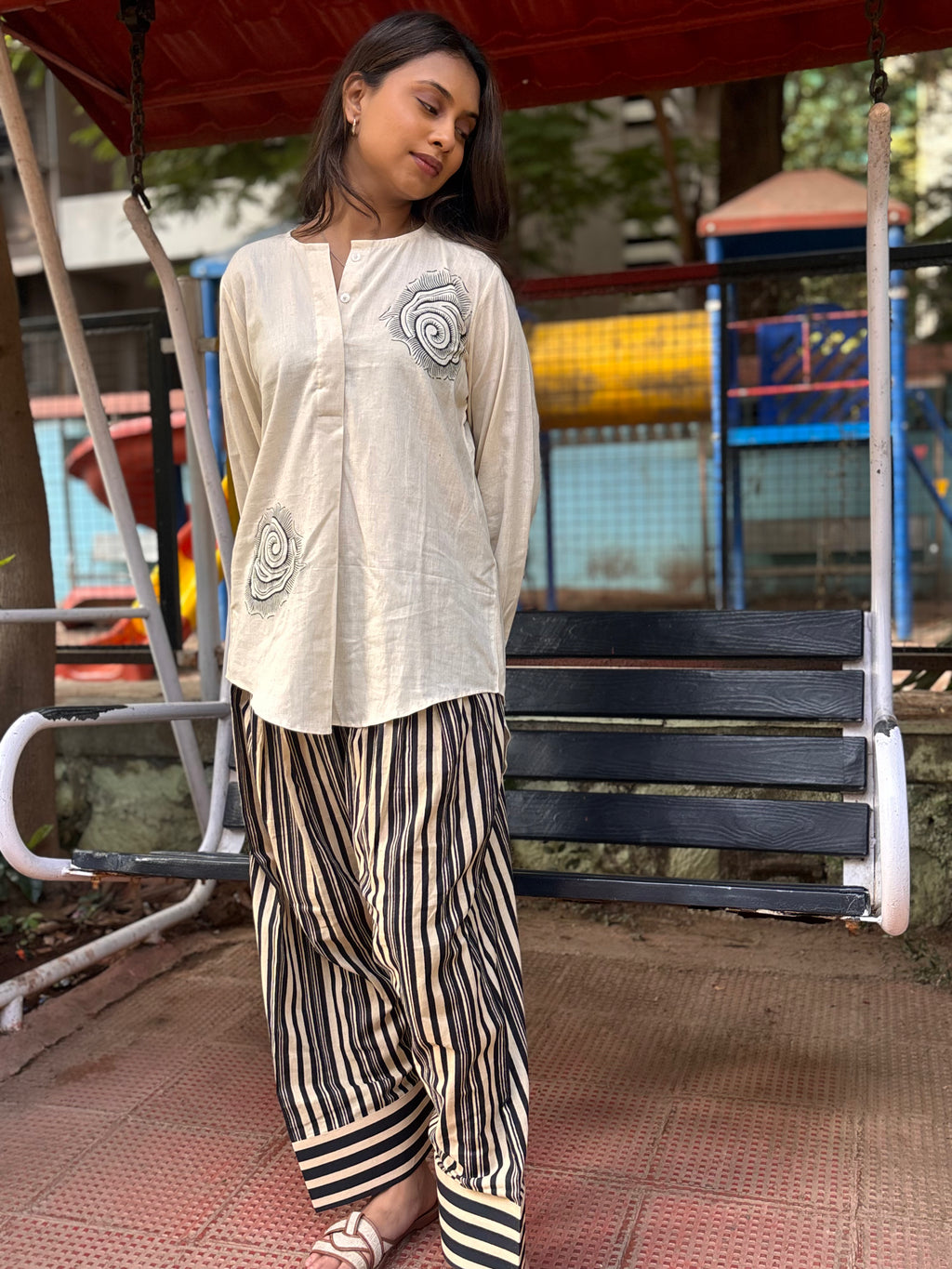 Woman wearing a white top with spiral designs and striped pants standing in front of a playground.