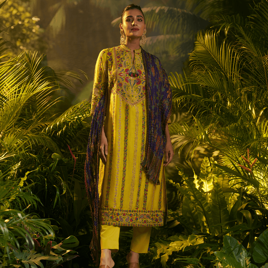 Model wearing a mustard yellow embroidered kurta set with intricate designs and a contrasting purple dupatta