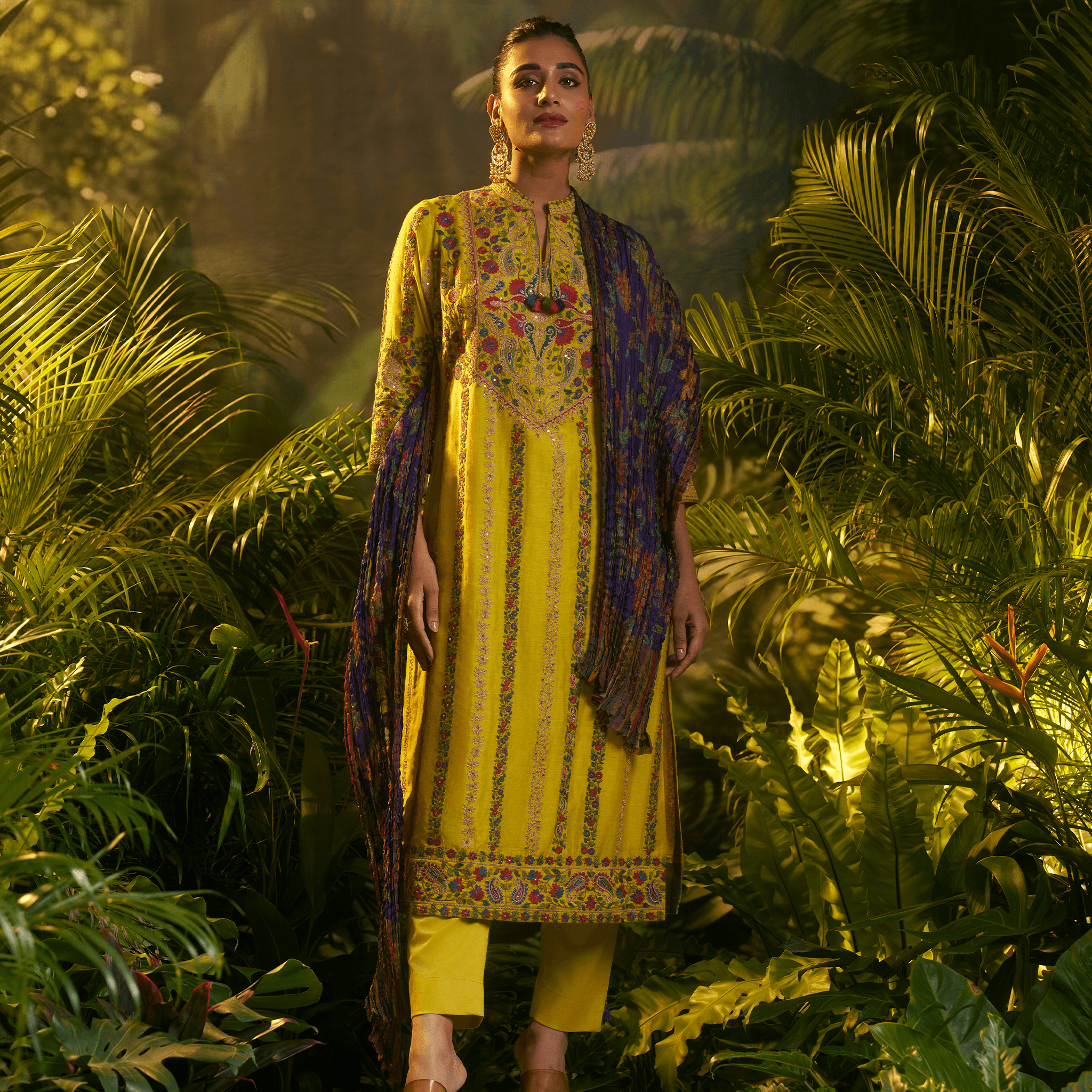 Model wearing a mustard yellow embroidered kurta set with intricate designs and a contrasting purple dupatta